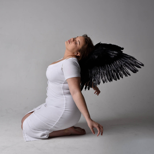 Full length portrait of young plus sized woman with short brunette hair,  wearing a tight white  dress and lack feather angel wings, kneeling pose on studio background. - Photo, Image