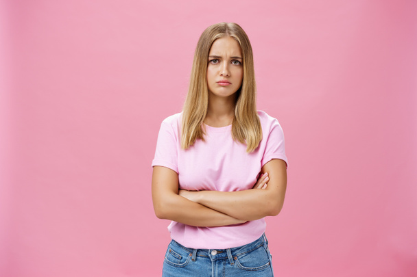 Sad envy and disappointed gloomy cute young woman with fair hair in casual t-shirt crossing arms against chest in upset gesture pursing lips and frowning looking concerned and sad over pink wall - Photo, Image