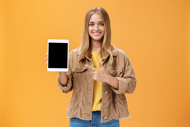 Get ready for autumn semister with new digital tablet. Portrait of charming female in corduroy jacket showing gadget screen pointing at device and smiling pleased and friendly over orange wall - Foto, immagini