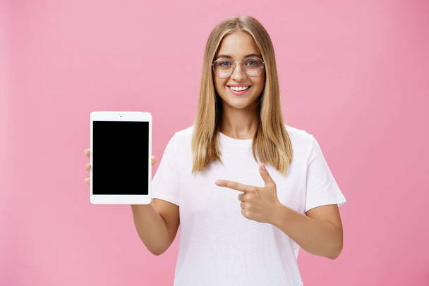 Στιγμιαία λήψη αισιόδοξης και χαρούμενης γυναίκας που δείχνει δροσερό ψηφιακό tablet να δείχνει την οθόνη της συσκευής και να χαμογελά ευρέως στην κάμερα δίνοντας συμβουλές ποια εφαρμογή είναι χρήσιμη ποζάροντας σε γυαλιά κατά του ροζ τοίχου - Φωτογραφία, εικόνα