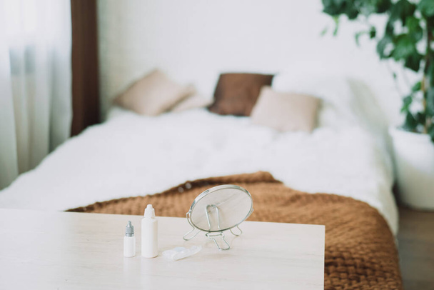 Eye drops and round mirror lie on the table in front of the bed. - Foto, Bild