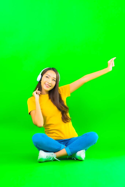 Retrato hermosa joven asiática mujer uso inteligente teléfono móvil con auriculares en verde aislado fondo - Foto, imagen
