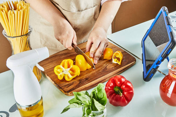Teenager prepares virtual online seminar, cuts yellow pepper, looks at digital recipe on touchscreen tablet while preparing healthy meal in the kitchen at home - Photo, Image