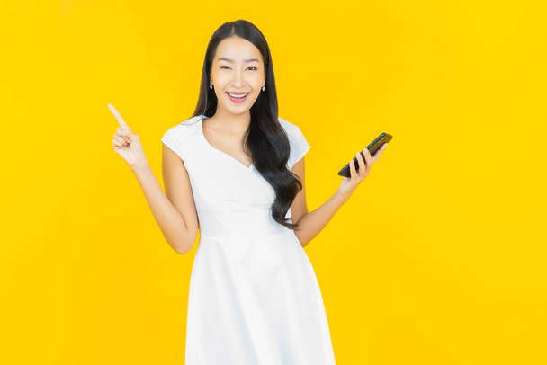 Muotokuva kaunis nuori aasialainen nainen hymyilee älypuhelimella keltaisella väritaustalla - Valokuva, kuva