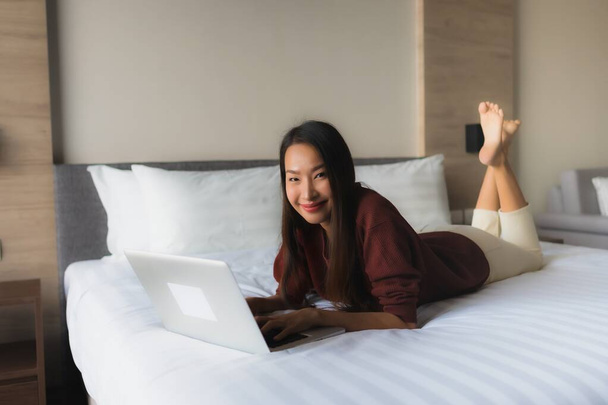 Portret piękne młode azjatyckie kobiety za pomocą komputera i telefonu komórkowego na łóżku we wnętrzu sypialni - Zdjęcie, obraz