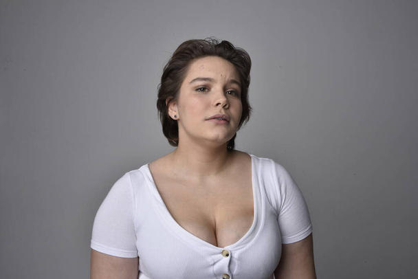 Retrato de cerca de una joven mujer de tamaño grande con pelo corto y morena, con una camisa blanca, con expresiones faciales emocionales sobre el fondo de un estudio de luz.   - Foto, Imagen