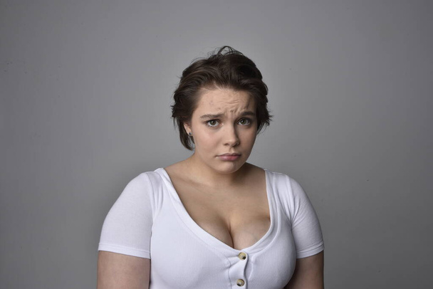 Close up portret młodej plus wielkości kobieta z krótkimi brunetkami włosy, noszenie białej koszuli, z ponad górnej emocjonalnej ekspresji twarzy na tle światła studio.   - Zdjęcie, obraz