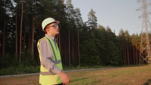 The young electrical engineer is tired of work, he throws up his helmet and walks away, he is wearing protective clothing and a hard hat. Concept. technology, green, electricity, tower - Footage, Video