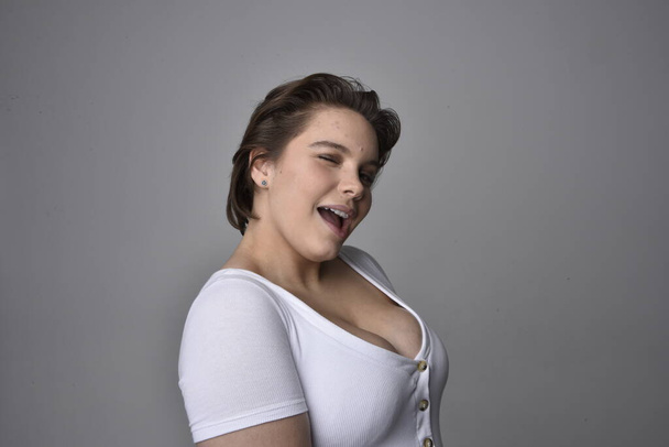 Close up portrait of young plus sized woman with short brunette hair,  wearing a white shirt, with over the top emotional facial expressions against a light studio background.   - Photo, Image