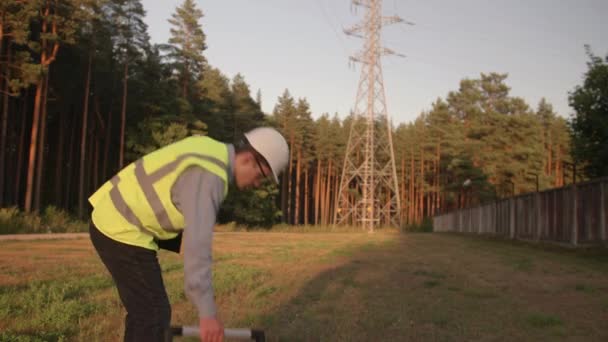 A young electrical engineer walks up to the electrical tower and looks around, wearing protective clothing and a hard hat. Concept. technology, green, electricity, tower - Footage, Video
