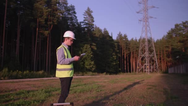 A young electrical engineer is reading something on a tablet, he is wearing protective clothing and a helmet. Concept. technology, green, electricity, tower - Footage, Video