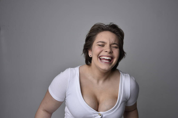 Close-up portret van jonge plus size vrouw met kort brunette haar, het dragen van een wit shirt, met over de top emotionele gezichtsuitdrukkingen tegen een lichte studio achtergrond.   - Foto, afbeelding
