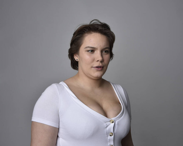 Close up portrait of young plus sized woman with short brunette hair,  wearing a white shirt, with over the top emotional facial expressions against a light studio background.   - Photo, Image