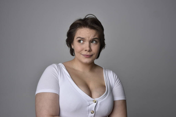 Κοντινό πορτραίτο νεαρής και μεγάλου μεγέθους γυναίκας με κοντά καστανά μαλλιά, φορώντας λευκό πουκάμισο, με πάνω από την κορυφή συναισθηματικές εκφράσεις του προσώπου σε ένα ελαφρύ στούντιο φόντο.   - Φωτογραφία, εικόνα