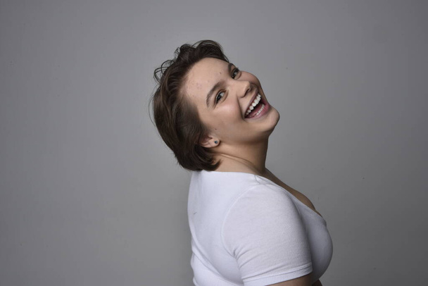 Close up portrait of young plus sized woman with short brunette hair,  wearing a white shirt, with over the top emotional facial expressions against a light studio background.   - Φωτογραφία, εικόνα