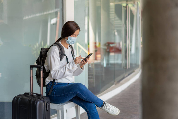 Reisen. Frau in weißem langärmeligem Hemd und Jeans mit Maske. auf dem Stuhl sitzen und Mobiltelefon mit Koffer auf der rechten Hand und Rucksack benutzen. - Foto, Bild