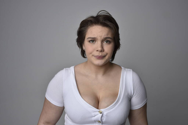 Κοντινό πορτραίτο νεαρής και μεγάλου μεγέθους γυναίκας με κοντά καστανά μαλλιά, φορώντας λευκό πουκάμισο, με πάνω από την κορυφή συναισθηματικές εκφράσεις του προσώπου σε ένα ελαφρύ στούντιο φόντο.   - Φωτογραφία, εικόνα
