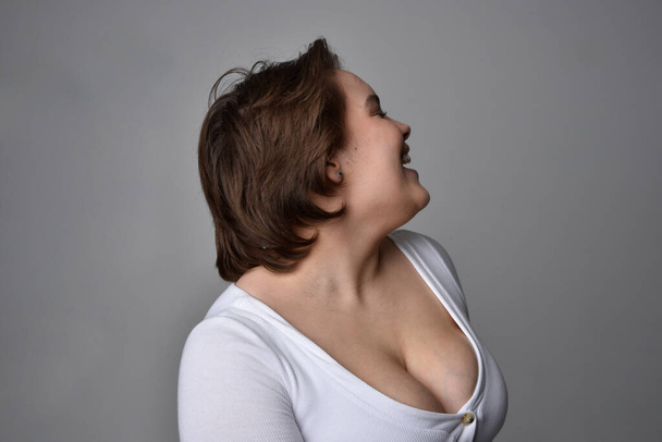 Retrato de cerca de una joven mujer de tamaño grande con pelo corto y morena, con una camisa blanca, con expresiones faciales emocionales sobre el fondo de un estudio de luz.   - Foto, Imagen