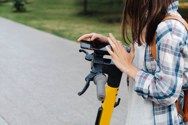 Close up image of woman hands paying for public hire electric scooter with a phone app. She's wearing checkered shirt. Cropped, half head. Next to a paved walk in the park. - 写真・画像