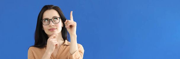 Pensive woman in glasses showing index finger up on blue background - Foto, imagen