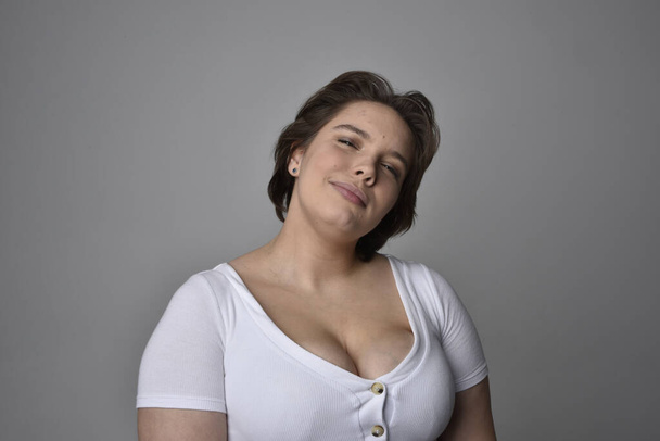 Retrato de cerca de una joven mujer de tamaño grande con pelo corto y morena, con una camisa blanca, con expresiones faciales emocionales sobre el fondo de un estudio de luz.   - Foto, imagen