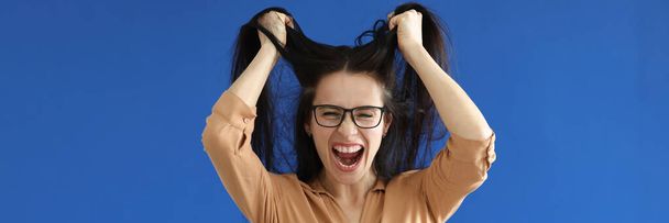 Mulher irritada com óculos rasgando seu cabelo no fundo azul - Foto, Imagem