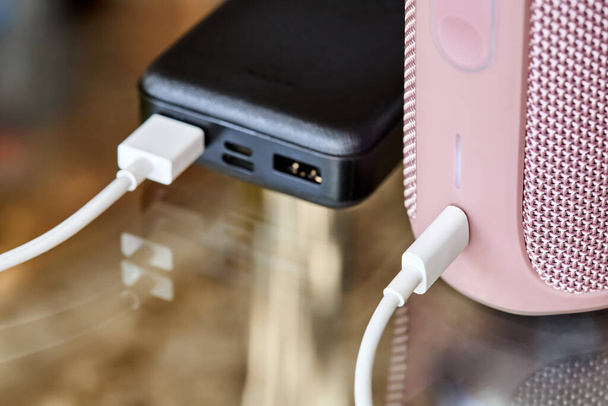 Power banka nabíjí přenosný audio reproduktor pomocí USB kabelu na skleněném povrchu. Detailní záběr, selektivní zaměření - Fotografie, Obrázek