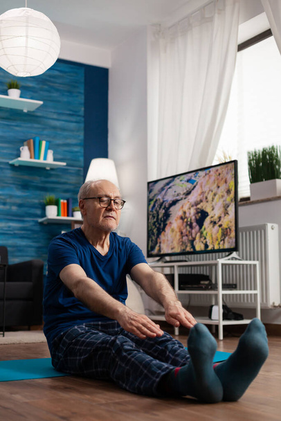 Összpontosított nyugdíjas visel sportruházat ül jóga szőnyegen nyújtózkodó lábak izom - Fotó, kép