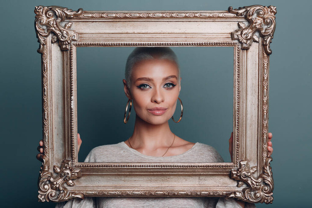 Millenial young woman with short blonde hair holds gilded picture frame in hands behind her face portrait. - Photo, Image