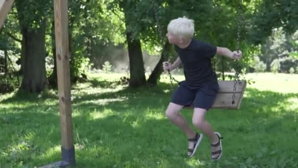 Un ragazzo albino cavalca un'altalena in estate in un parco ombreggiato - Filmati, video