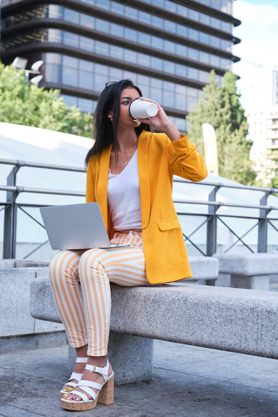 Giovane donna d'affari latina con giacca gialla che lavora con un computer portatile e beve caffè seduta all'aperto. - Foto, immagini