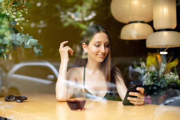 aantrekkelijk meisje met behulp van de telefoon tijdens het zitten in een modern café en drinken. uitzicht vanaf de straat door glas - Foto, afbeelding