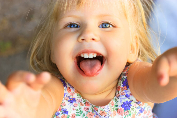 Little blonde preschool girl reaches up with both hands, looks at a camera. A blue-eyed cute female child 2-3 years old smiles with open mouth and tongue sticking out. A pretty playful kid. Pick me up - Photo, Image