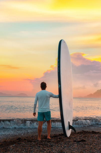 Fitter Mann mit Surfbrett und posiert am Strand. Im Hintergrund das Meer und der Sonnenuntergang. Vertikal. Kopierraum. Sommer-Surfen und extreme Aktivität. - Foto, Bild