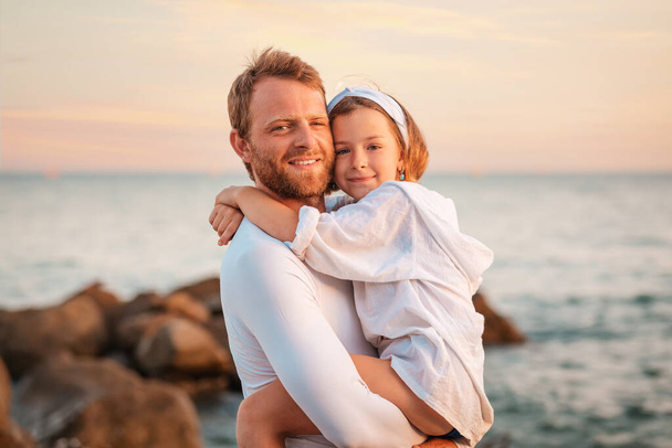Dzień Ojca. Portret uśmiechniętego ojca przytulającego swoją przedszkolną córkę w ramionach. W tle, morze i niebo. Koncepcja szczęśliwego ojcostwa i adopcji dzieci. - Zdjęcie, obraz