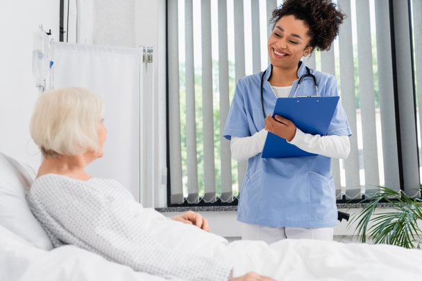 Cheerful african american nurse with clipboard looking at senior patient on bed  - Фото, зображення