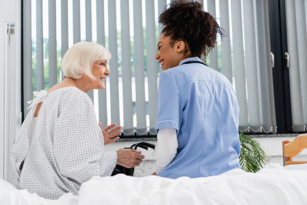 Side view of cheerful senior woman talking to african american nurse on bed  - Foto, Bild