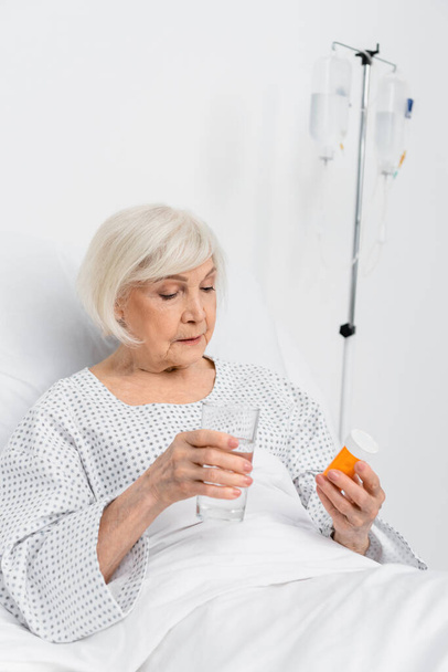 Elderly patient holding pills and glass of water in hospital ward  - Foto, afbeelding