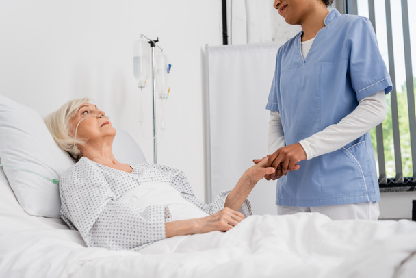Afrikai-amerikai nővér fogja a kezét vezető beteg orrkanül kórházi ágyon  - Fotó, kép