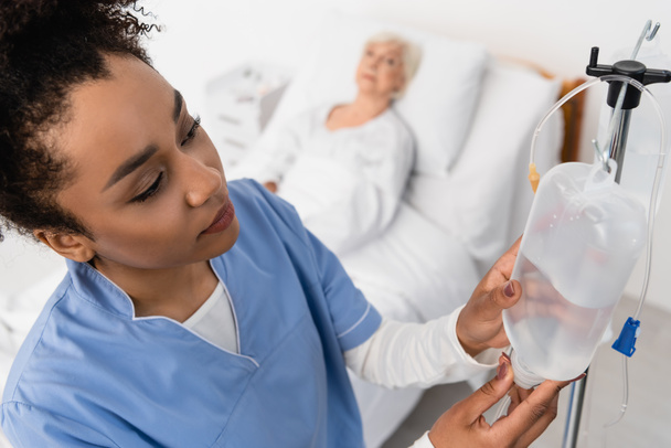 Vista ad alto angolo dell'infermiera afro-americana che tiene una bottiglia per la terapia endovenosa vicino al paziente offuscato  - Foto, immagini