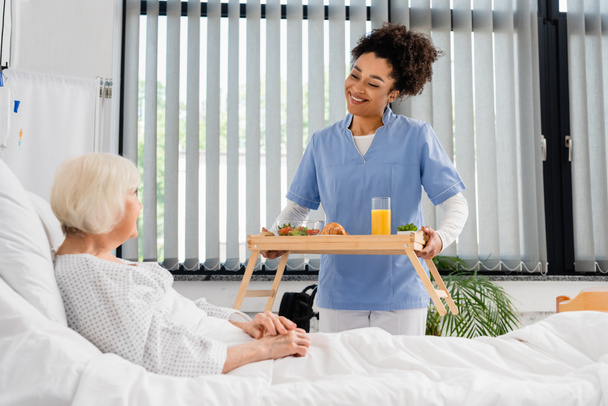 Enfermera afroamericana sonriente sosteniendo desayuno y jugo de naranja en bandeja cerca del paciente en el hospital  - Foto, Imagen