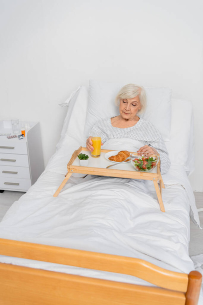 Elderly patient holding glass of orange juice near food in hospital ward  - 写真・画像