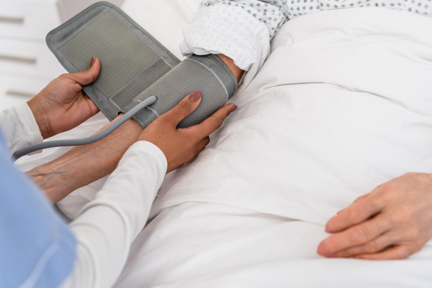 Ausgeschnittene Ansicht einer afrikanisch-amerikanischen Krankenschwester mit Tonometer an der Hand eines älteren Patienten auf dem Krankenhausbett  - Foto, Bild