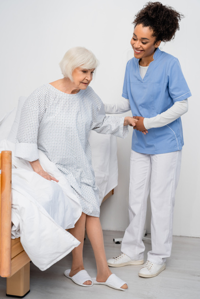 Smiling african american nurse holding hand of senior patient in hospital ward  - Photo, Image