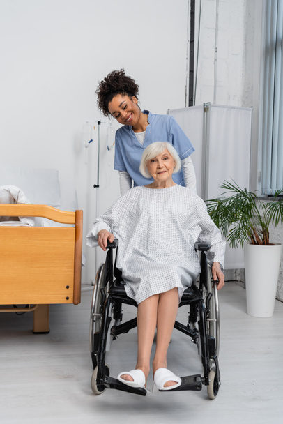 African american nurse smiling near senior patient in wheelchair in clinic  - Zdjęcie, obraz