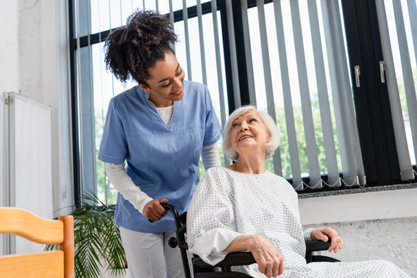 Smiling african american nurse looking at senior patient in wheelchair in hospital  - Zdjęcie, obraz