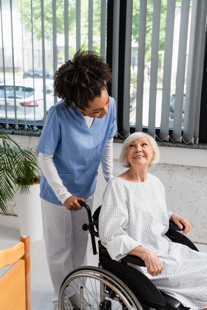 Afrykańska pielęgniarka amerykańska patrzy na starszego pacjenta w sukni i wózku inwalidzkim  - Zdjęcie, obraz