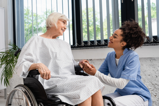 Afričanky americká zdravotní sestra drží ruku usměvavé starší ženy na invalidním vozíku  - Fotografie, Obrázek
