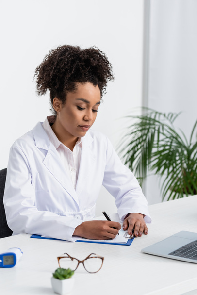 African american doctor in white coat writing on clipboard near laptop and pyrometer  - 写真・画像