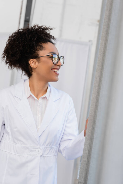Side view of cheerful african american doctor standing near curtains  - Photo, Image